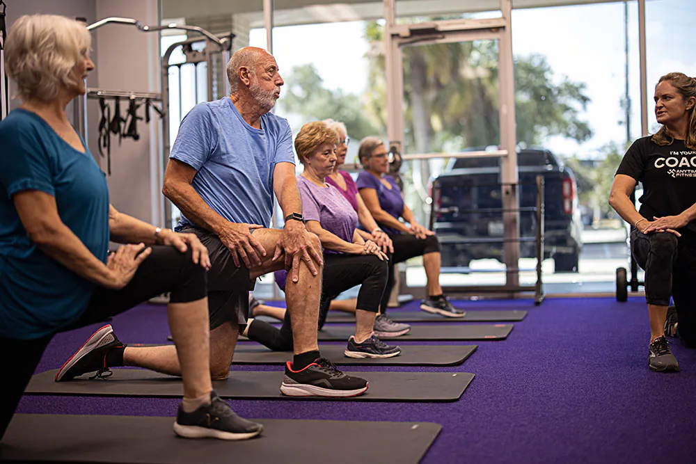 A man performs dips on a bench while a fitness coach kneels beside him, offering guidance in a gym setting filled with equipment.