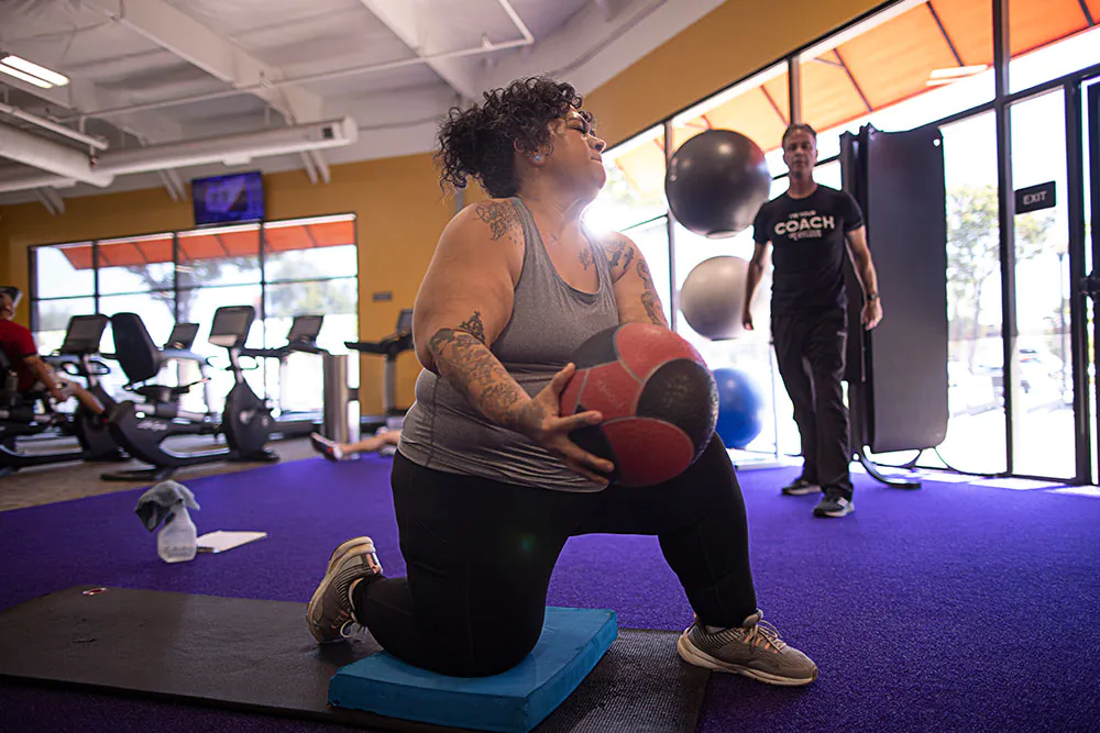 A man performs dips on a bench while a fitness coach kneels beside him, offering guidance in a gym setting filled with equipment.