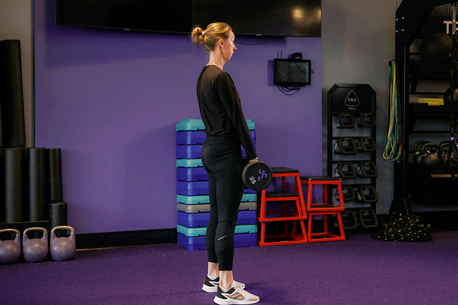 A woman in a gym performing a dumbbell Romanian deadlift. She stands with a neutral spine, hinges at her hips to lower a dumbbell toward her feet while keeping her legs relatively straight, and then returns to a standing position.