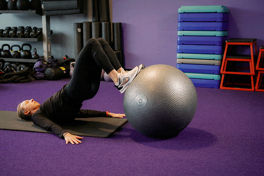 A woman lying on her back on a gym mat performing a stability ball hamstring curl. Her heels are placed on top of a large silver exercise ball; she lifts her hips into a bridge position and rolls the ball toward her glutes by bending her knees, then rolls it back out to the starting position.