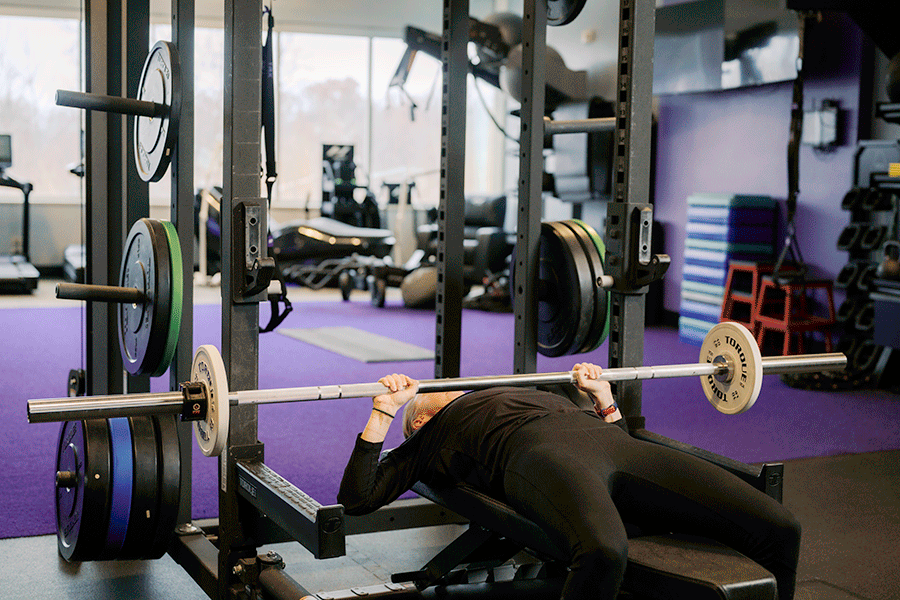 A woman performing a barbell chest press on a flat bench inside a power rack. She unearths the barbell with a wide grip, lowers it slowly to her mid-chest, and then presses it back up to the starting position with her arms fully extended.