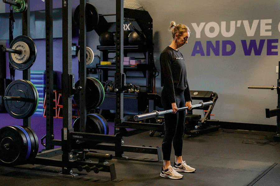 A woman performing a barbell hang clean in a gym. Starting with the barbell at mid-thigh height, she uses an explosive hip hinge and shrug to pull the bar upward, quickly rotating her elbows underneath to catch the barbell across the front of her shoulders in a partial squat position.