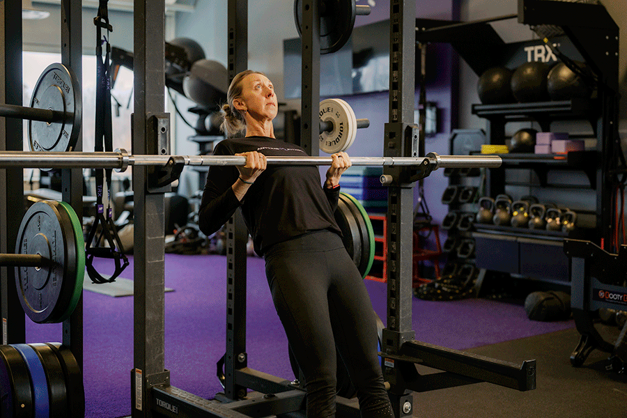 A woman in a gym performing a modified self-row (inverted row) using a barbell secured in a power rack. She hangs underneath the bar with her body in a straight line and her feet planted on the floor, then pulls her chest up toward the bar by driving her elbows back before lowering herself with control.