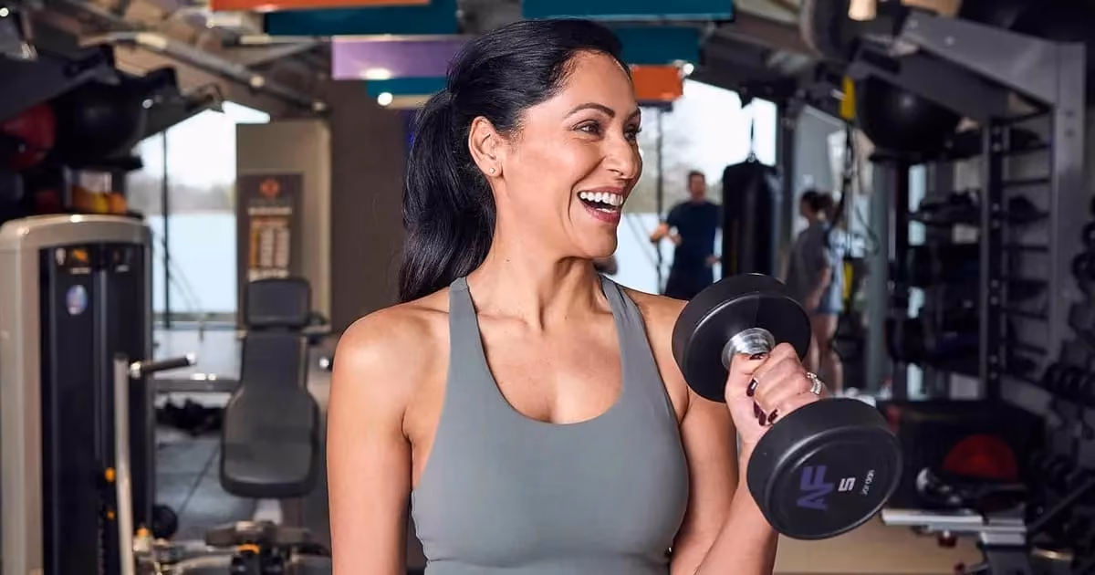 A woman in a gym lifts a dumbbell while smiling, wearing a gray tank top. She appears energetic and confident. 