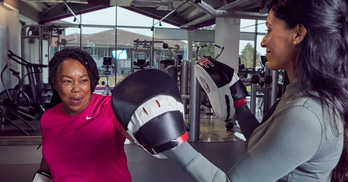 Two women training in a gym. One wears a pink shirt and punches a black focus mitt held by the other, who wears a gray top. 