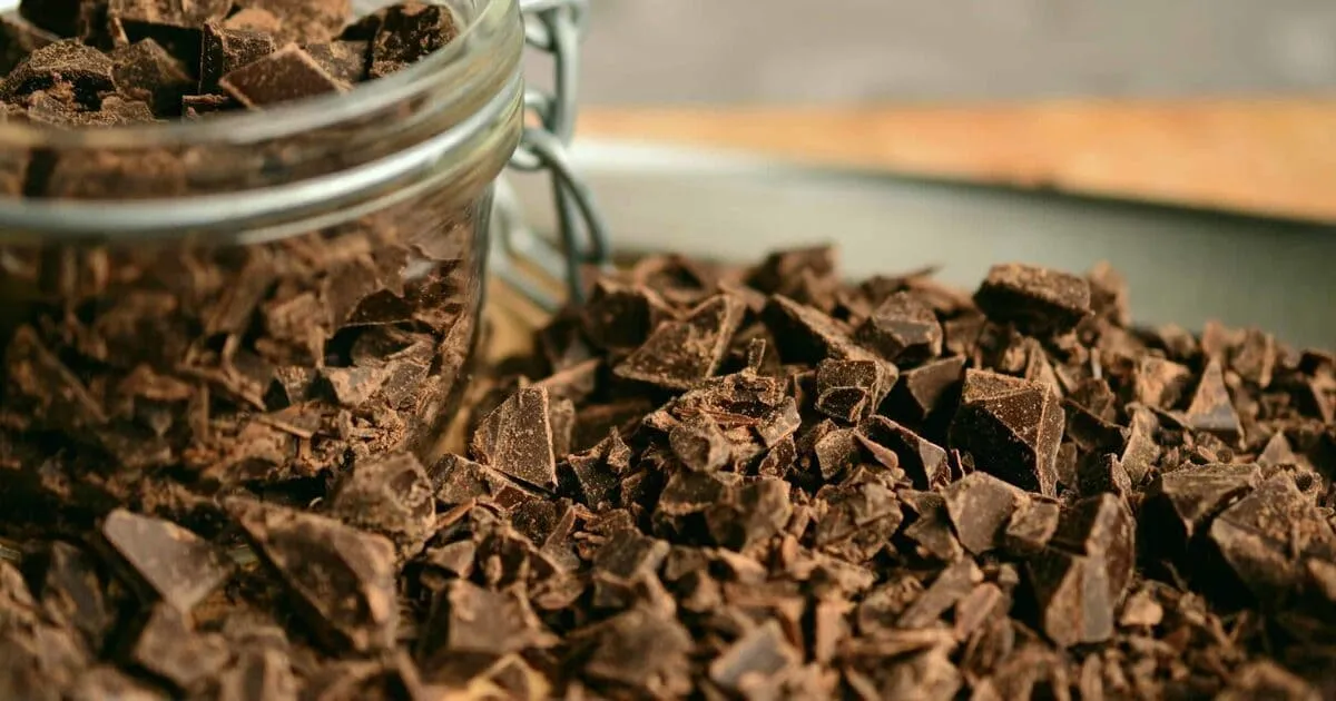 Close-up of dark chocolate chunks spilling from an open glass jar onto a wooden surface, creating a tempting and indulgent atmosphere.
