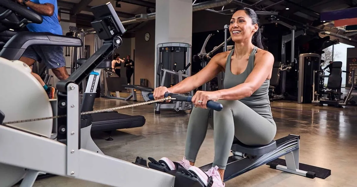 A woman in activewear is smiling while using a rowing machine at a gym. 
