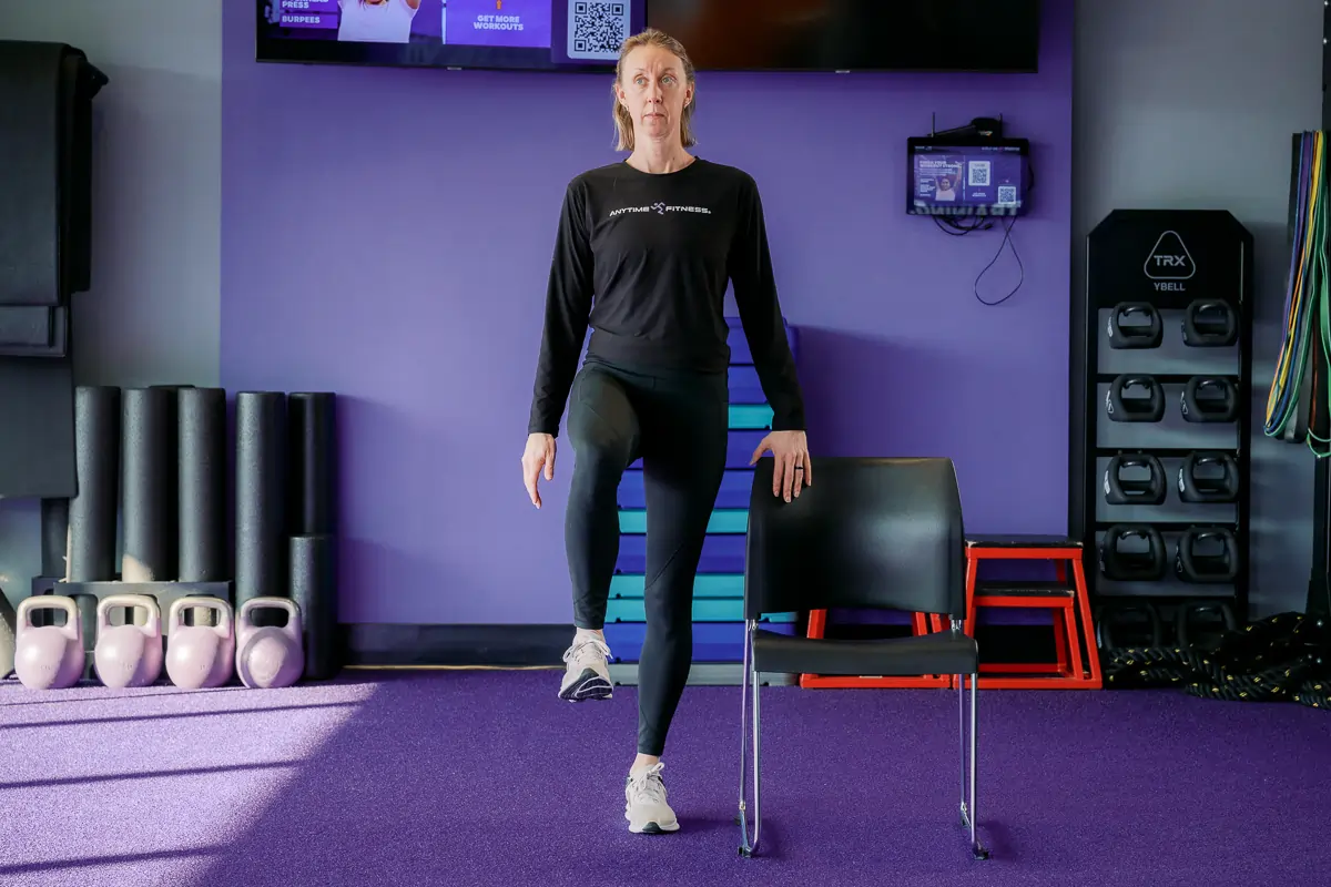 Anytime Fitness Coach demonstrating a supported single-leg stand, a strength and balance exercise.