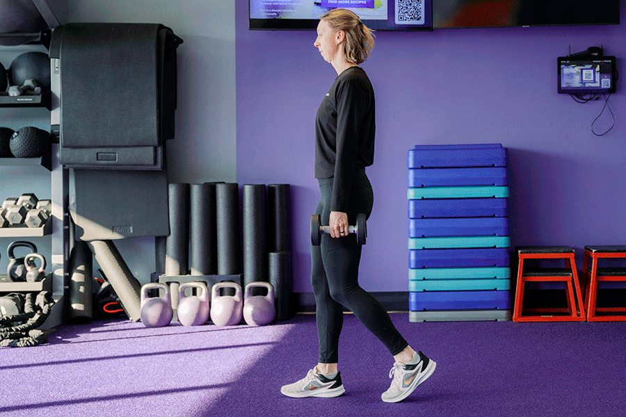 Anytime Fitness Coach demonstrating a farmer carry with dumbbells, a strength and balance exercise.