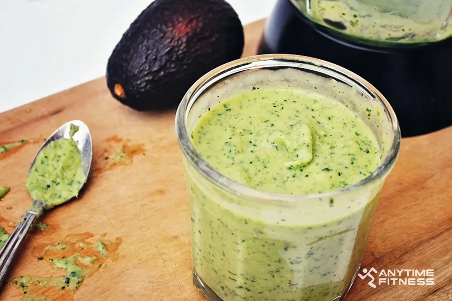 A close-up of a glass jar filled with a green avocado pre-workout smoothie sitting on a wooden cutting board, with a silver spoon, a whole ripe avocado and part of a black blender visible in the background.