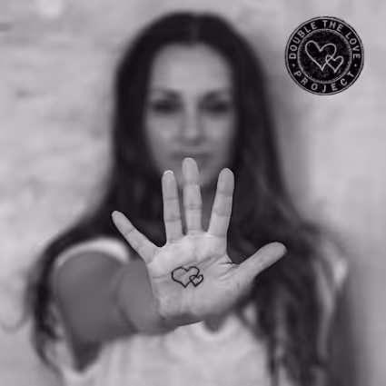 Black and white photo of a woman holding her hand forward with a heart symbol drawn on her palm, featuring the “Double the Love Project” logo.