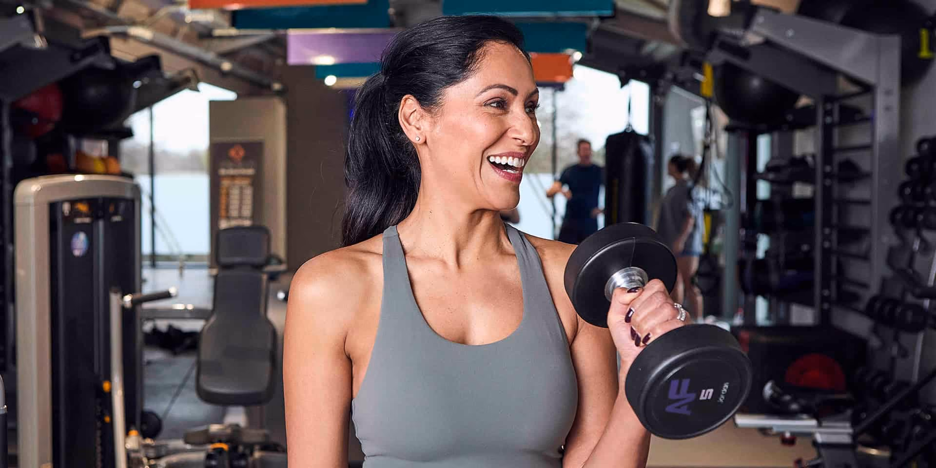 A woman in a gym holds a dumbbell, smiling confidently. She wears a gray tank top.
