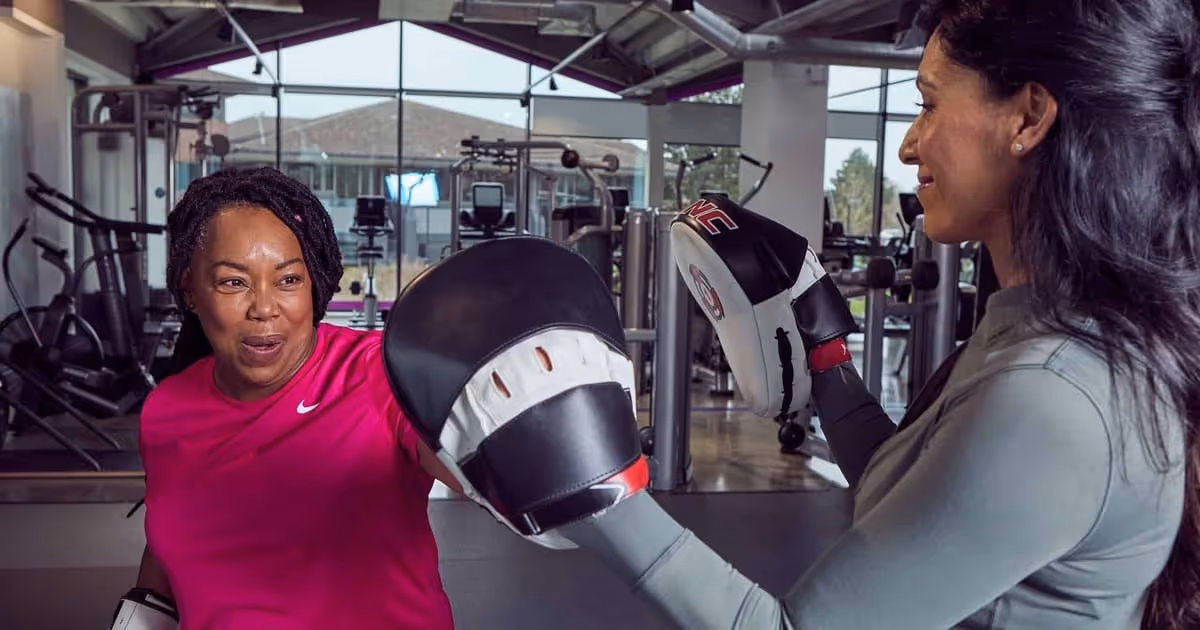 Two women training in a gym. One wears a pink shirt and punches a black focus mitt held by the other, who wears a gray top.