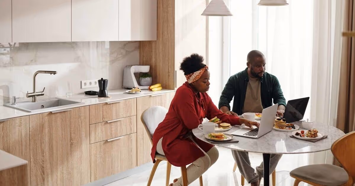 A couple sits at a round kitchen table with breakfast dishes and a laptop. The kitchen is modern with light wood cabinets.