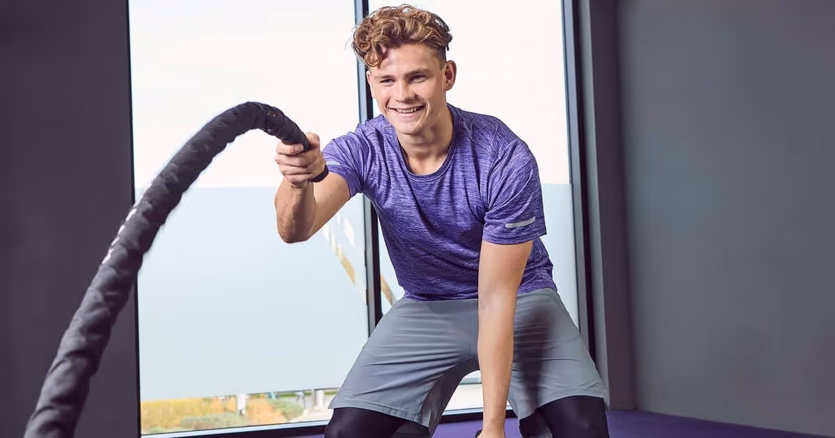 Young man exercises with battle ropes in a gym, wearing a purple shirt and gray shorts. He smiles energetically, conveying determination and joy.