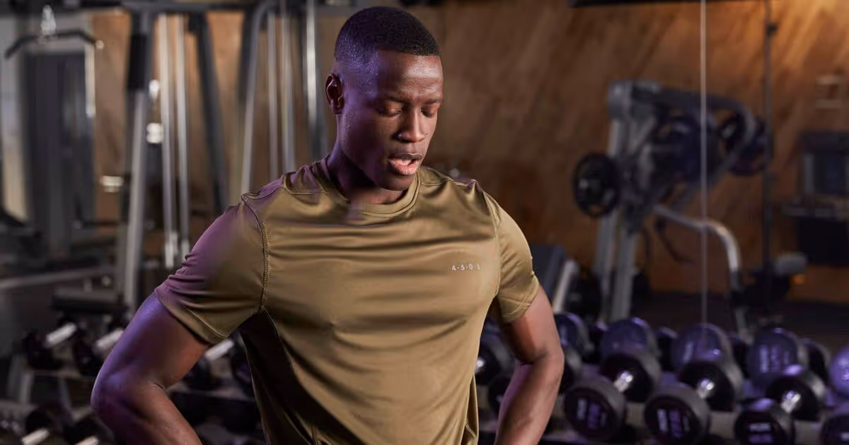 A man in an olive green workout shirt stands with hands on hips in a gym.