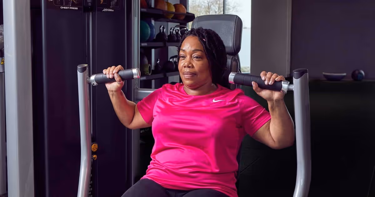 A woman in a bright pink shirt uses a chest press machine at the gym, focused and determined.