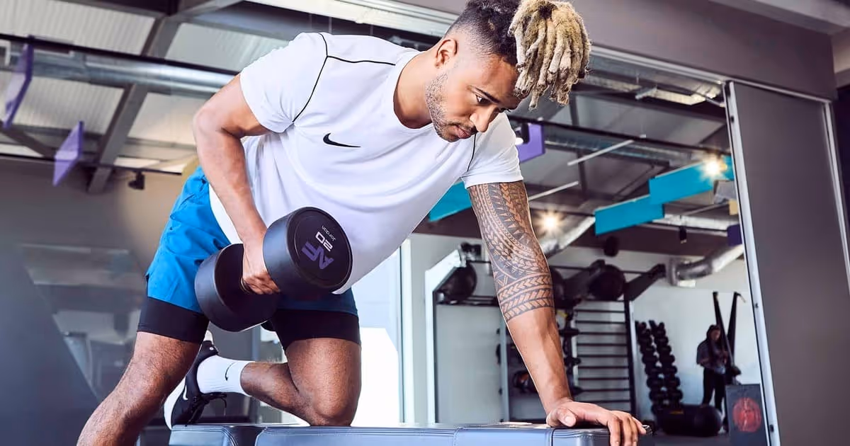 A man in a gym does a one-arm dumbbell row on a bench. He wears a white shirt and blue shorts, focusing intently.