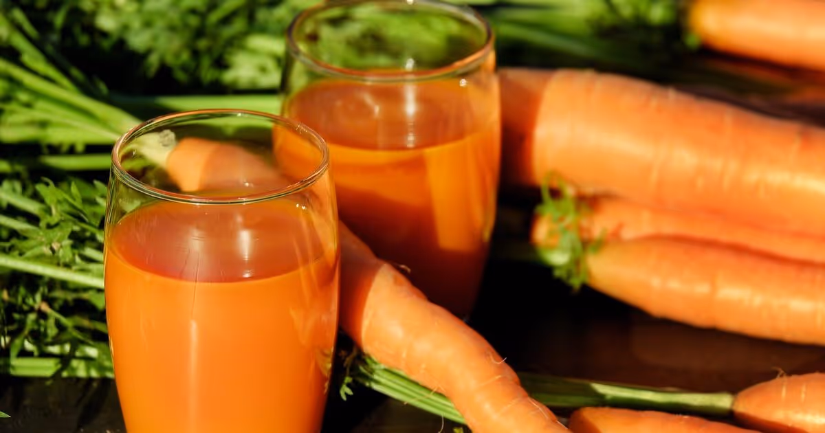 Two glasses of vibrant orange carrot juice are surrounded by fresh, whole carrots with green tops, creating a fresh and healthy atmosphere.
