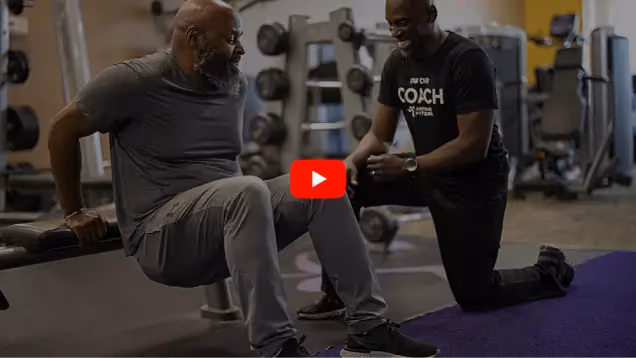 A man performs dips on a bench while a fitness coach kneels beside him, offering guidance in a gym setting filled with equipment.
