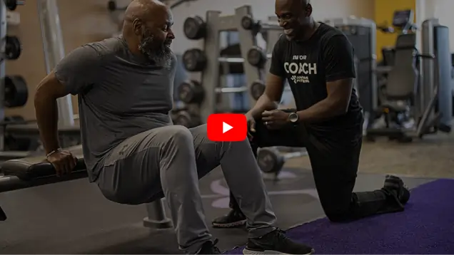 A man performs dips on a bench while a fitness coach kneels beside him, offering guidance in a gym setting filled with equipment.