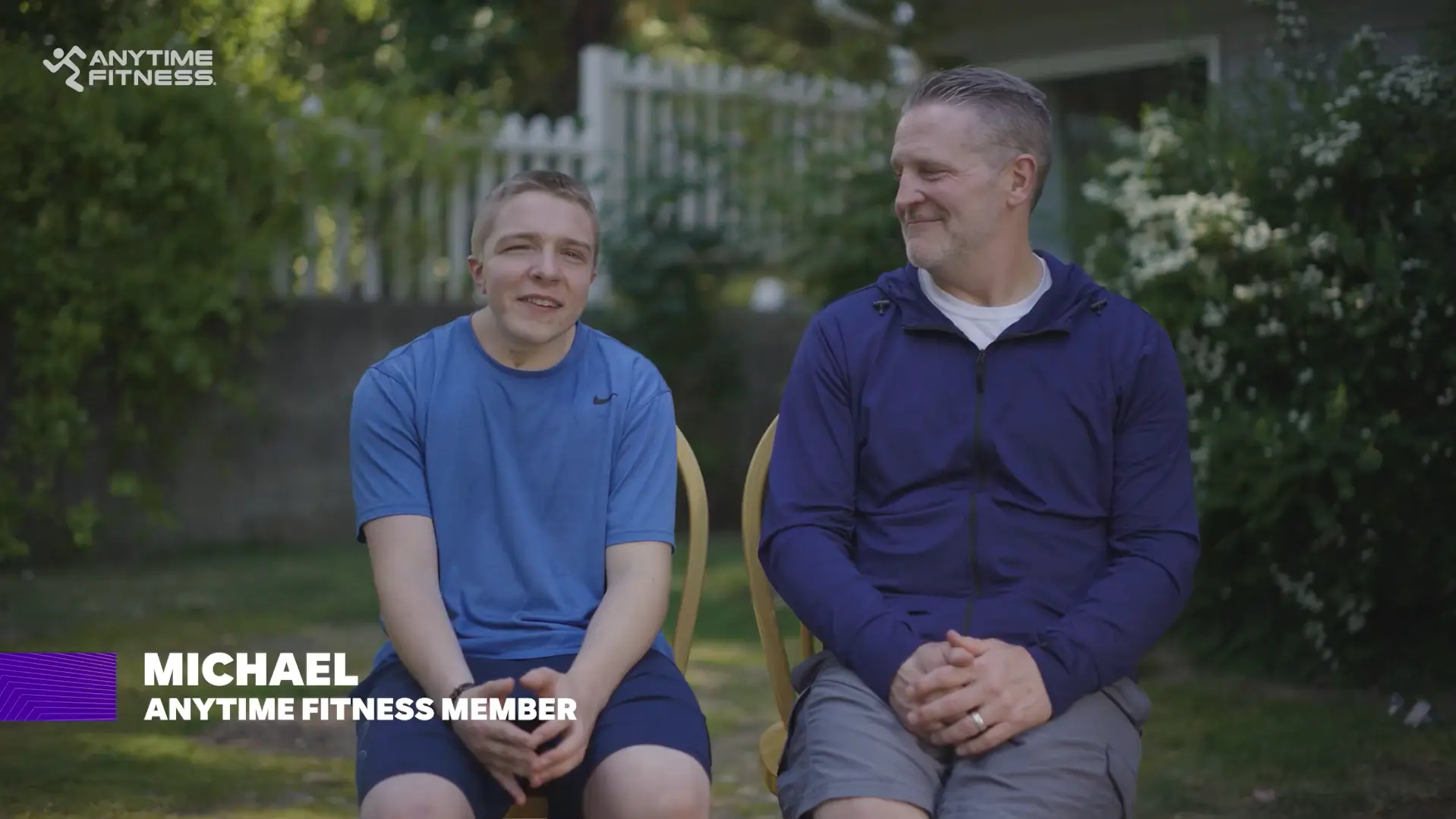A teenage boy and an older man sit side by side on chairs in a backyard, smiling during an interview. On-screen text identifies the boy as ‘Michael, Anytime Fitness Member.’