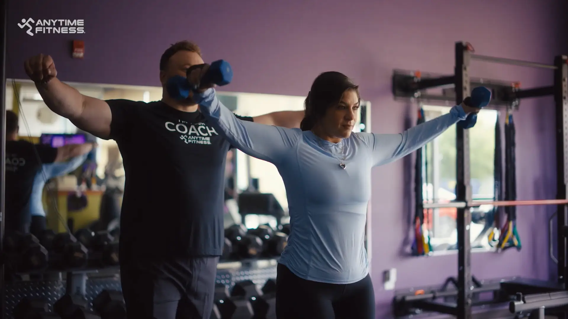 A personal trainer stands behind a woman in a gym, guiding her as she performs a shoulder raise with blue dumbbells. Strength equipment and mirrors are visible in the background.