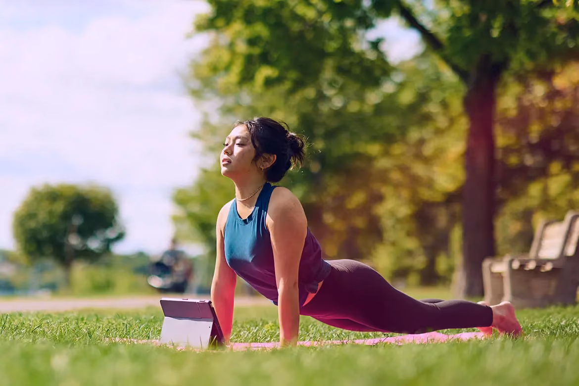 A woman practicing a Cobra yoga pose on a pink mat in a sunlit park, using a tablet to follow a guided session, illustrating a peaceful and accessible introduction to yoga.