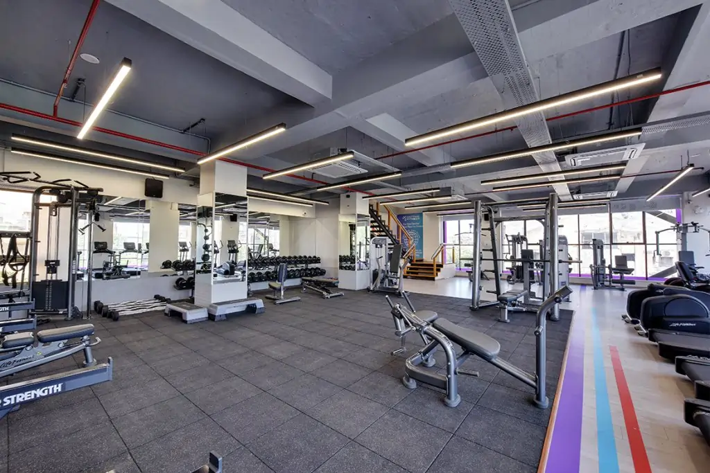 A wide-angle view of a modern, well-equipped Anytime Fitness gym floor featuring strength training machines, a dumbbell rack, power racks, and cardio equipment in a clean, professional setting.