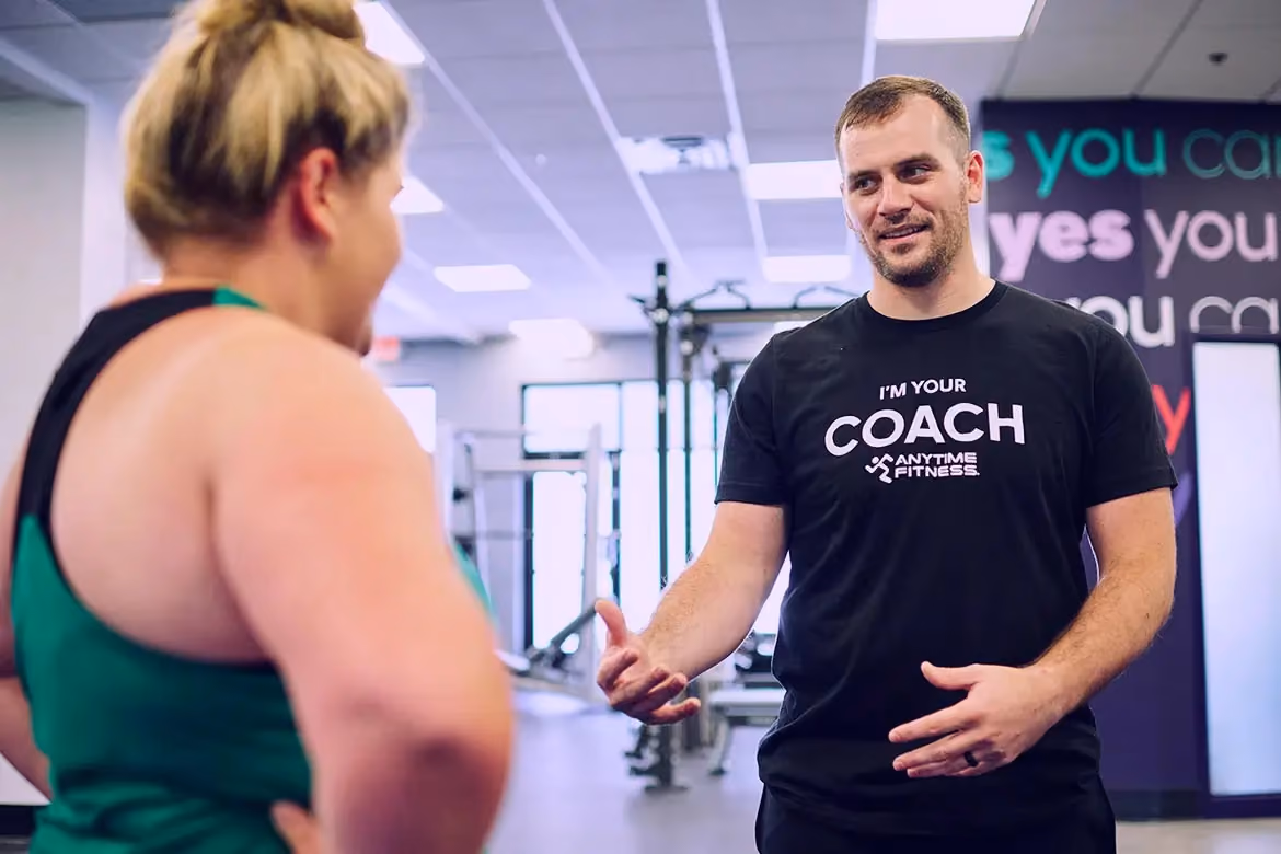 A supportive Anytime Fitness coach in a black "I'm Your Coach" t-shirt talking to a female gym member during a workout session, highlighting a welcoming and professional gym environment.