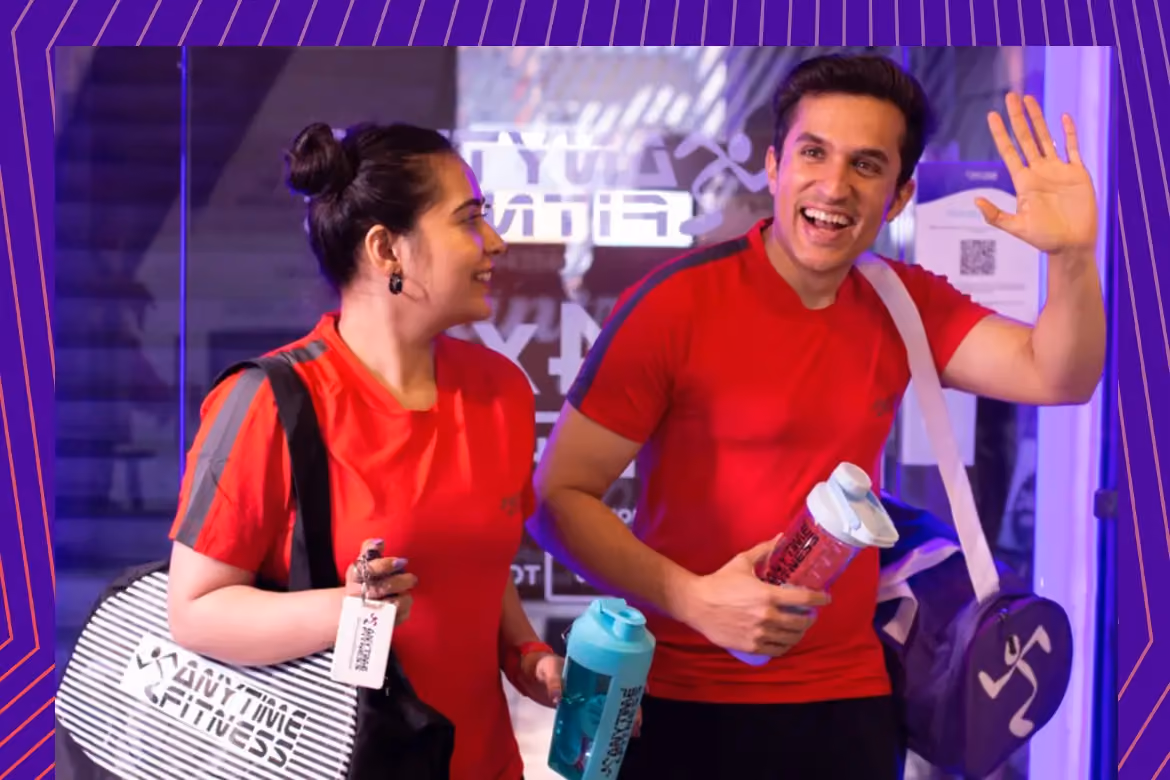 Two happy gym members in red Anytime Fitness shirts carrying gym bags and protein shakers while leaving the gym, illustrating the fitness lifestyle and post-workout recovery.