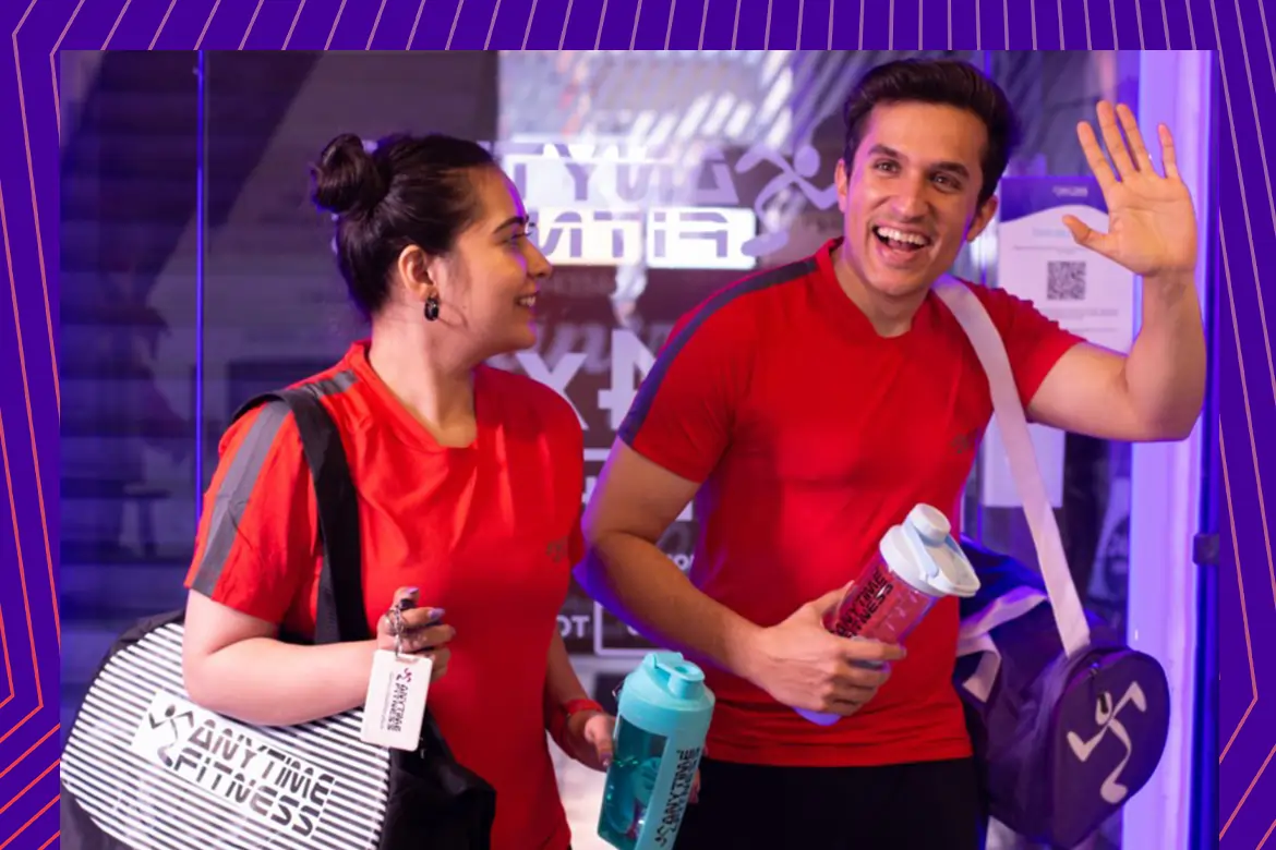 Two happy gym members in red Anytime Fitness shirts carrying gym bags and protein shakers while leaving the gym, illustrating the fitness lifestyle and post-workout recovery.