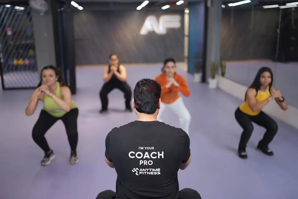 A back view of an Anytime Fitness Coach Pro leading a small group of four people through a squat exercise in a modern purple-floored gym studio.