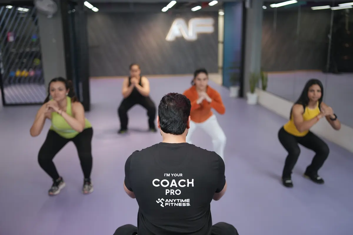 A back view of an Anytime Fitness Coach Pro leading a small group of four people through a squat exercise in a modern purple-floored gym studio.