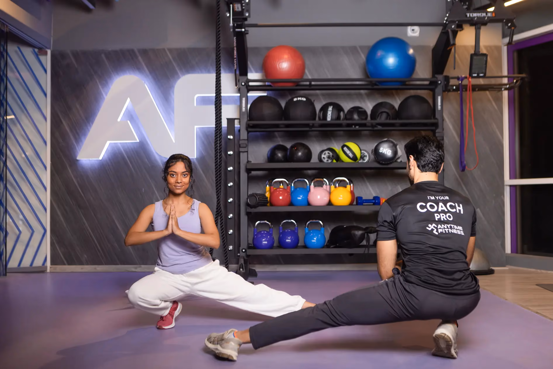 An Anytime Fitness personal trainer providing expert form guidance to a woman preparing to perform a barbell deadlift in the weightlifting area.