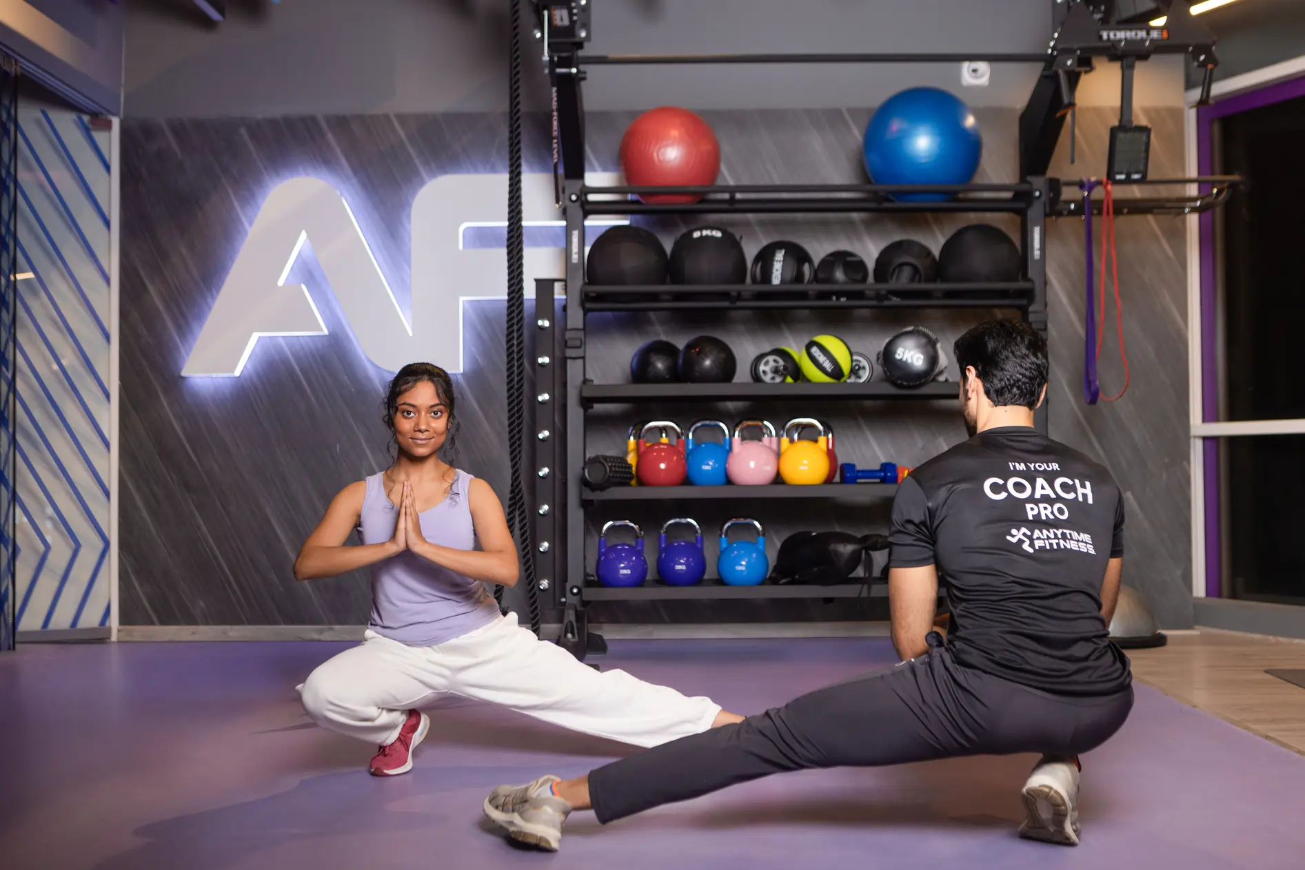 An Anytime Fitness personal trainer providing expert form guidance to a woman preparing to perform a barbell deadlift in the weightlifting area.