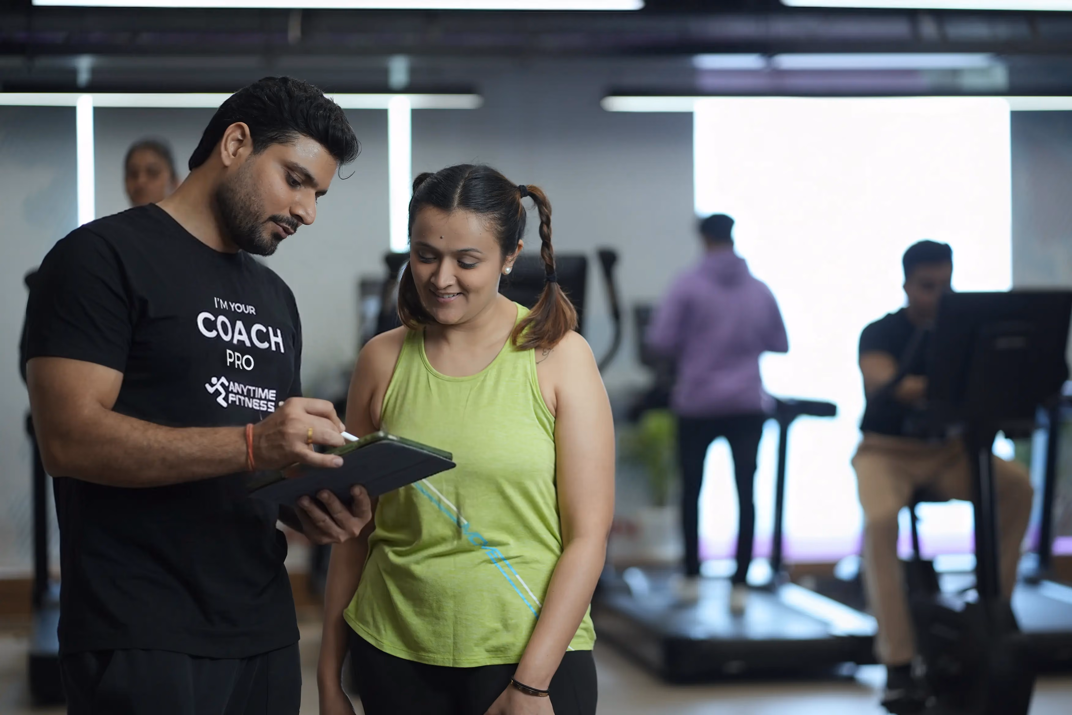 An Anytime Fitness Coach Pro using a digital tablet to discuss a personalized workout plan with a female member during a fitness consultation.