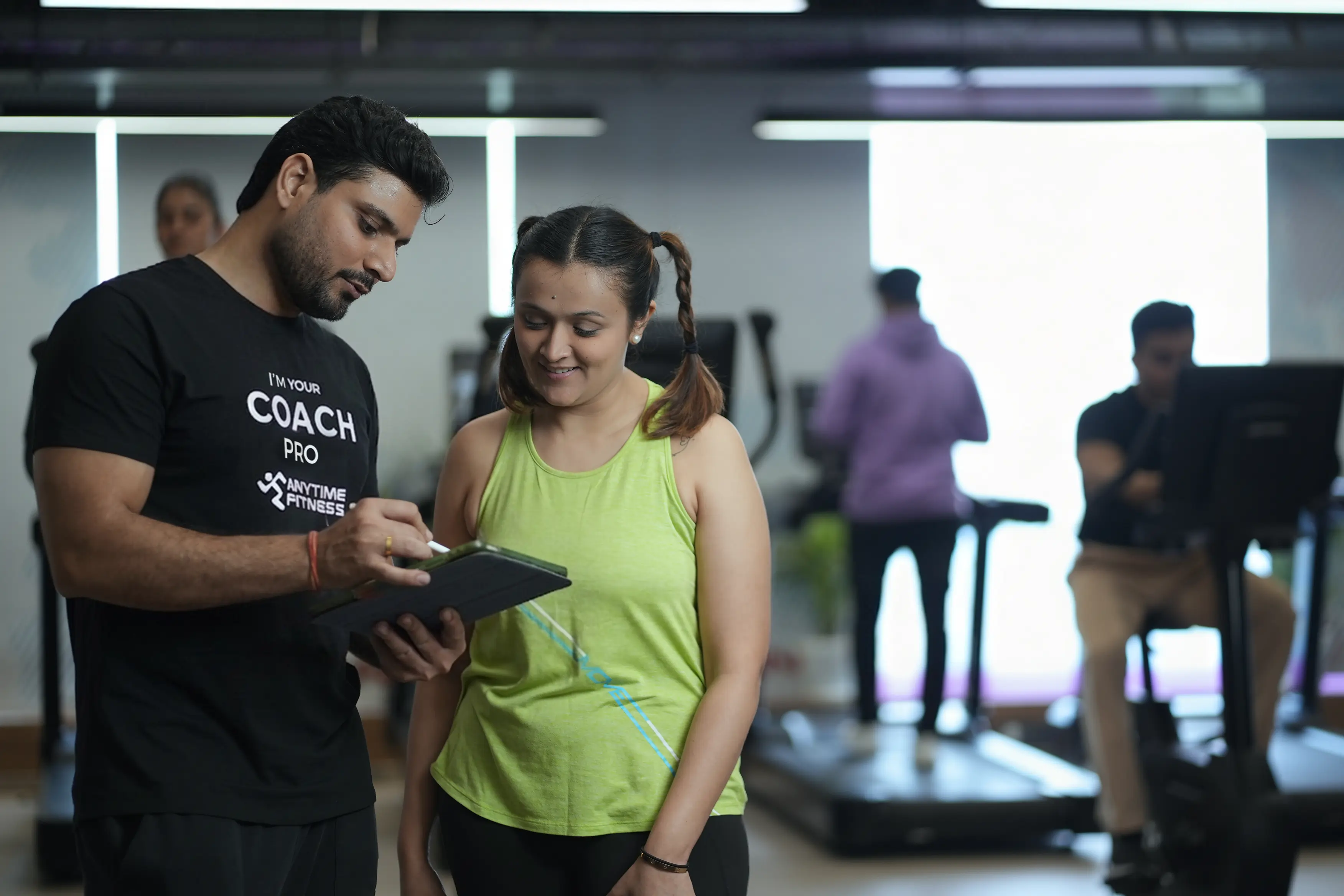 An Anytime Fitness Coach Pro using a digital tablet to discuss a personalized workout plan with a female member during a fitness consultation.