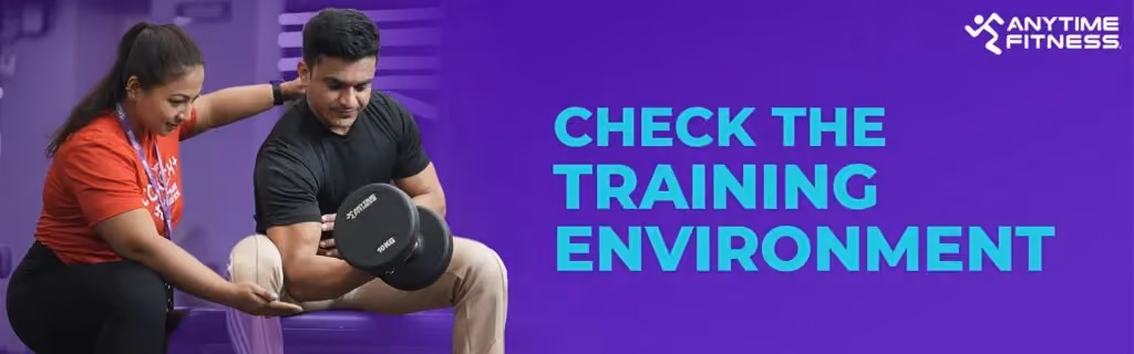 A banner titled "CHECK THE TRAINING ENVIRONMENT" featuring a female coach in a red shirt guiding a male member as he performs bicep curls with a dumbbell.