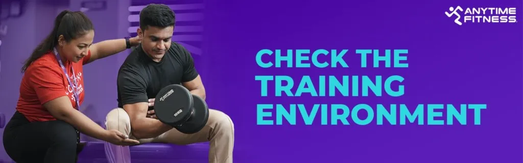 A banner titled "CHECK THE TRAINING ENVIRONMENT" featuring a female coach in a red shirt guiding a male member as he performs bicep curls with a dumbbell.