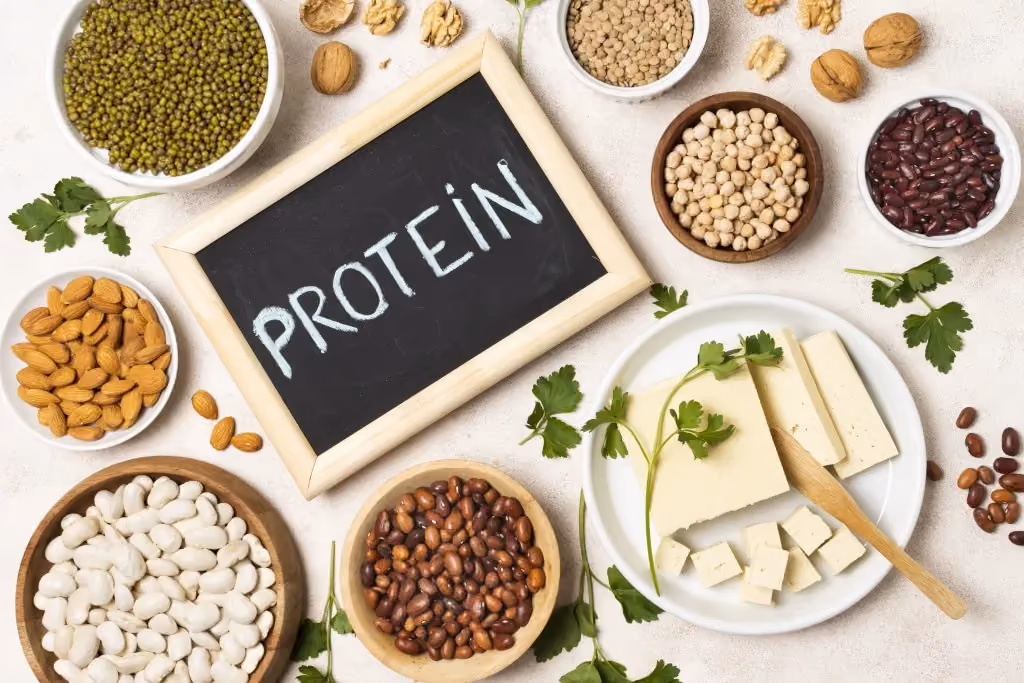 A flat-lay arrangement of plant-based protein sources surrounding a small chalkboard that says "PROTEIN." The image includes bowls of almonds, mung beans, chickpeas, kidney beans, white beans, walnuts, and a plate of sliced tofu garnished with parsley.