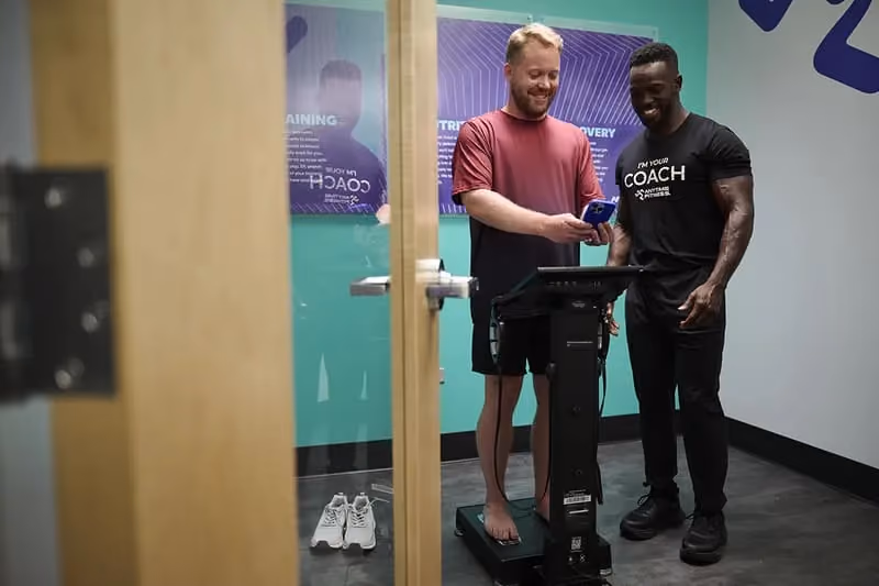 Two men in a gym room, one on a body composition scale holding a device, the other wearing a "Coach" shirt assisting him.