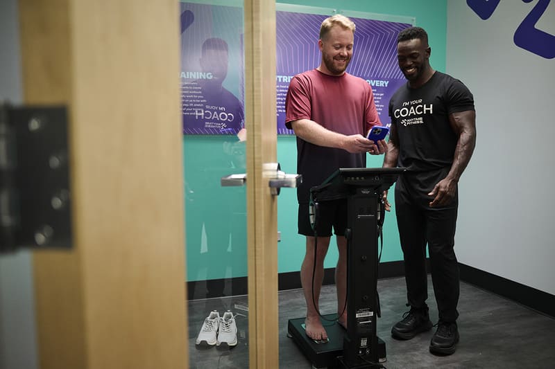 Two men in a gym room, one on a body composition scale holding a device, the other wearing a "Coach" shirt assisting him.