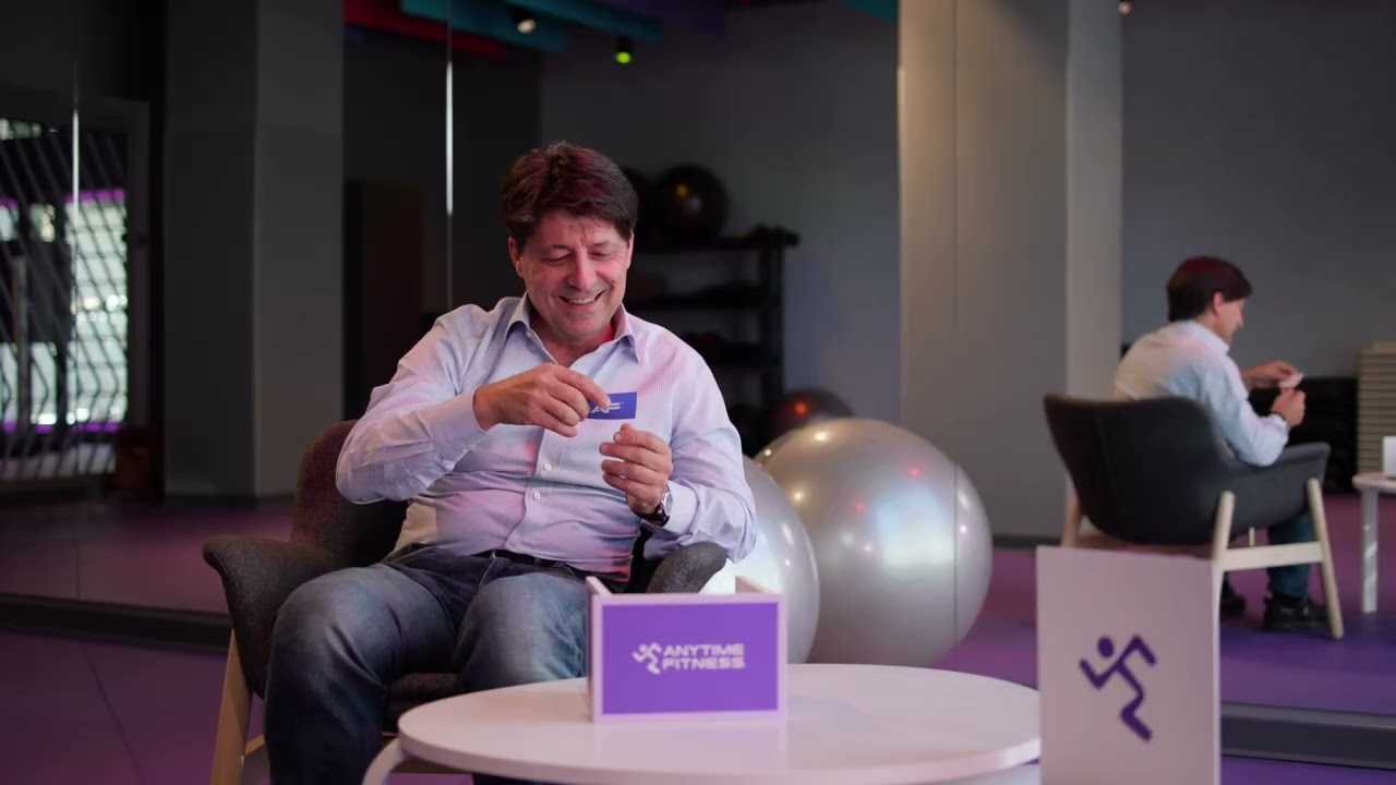 Ein Mann im gestreiften Hemd sitzt während eines Interviews in einem grauen Sessel in einem Anytime Fitness Studio. Im Hintergrund sind Gymnastikbälle, Trainingsgeräte und Zimmerpflanzen zu sehen.