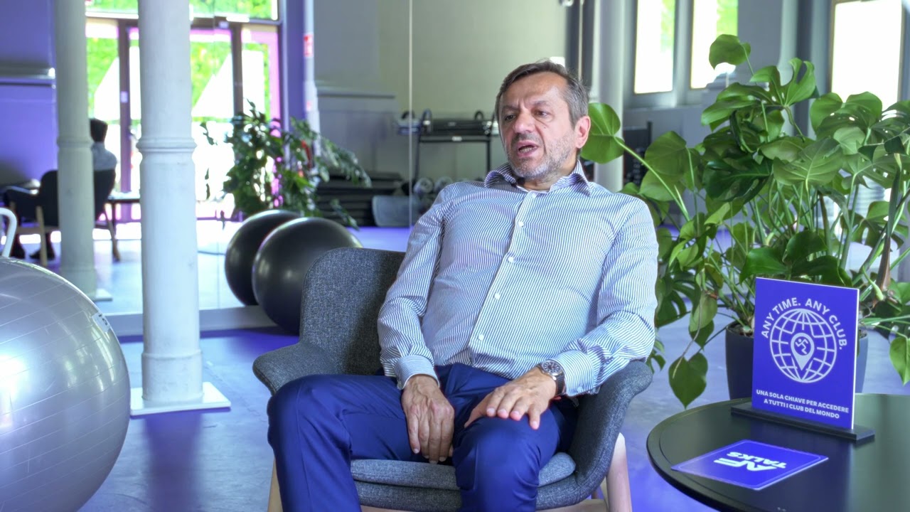 Ein Mann im gestreiften Hemd sitzt während eines Interviews in einem grauen Sessel in einem Anytime Fitness Studio. Im Hintergrund sind Gymnastikbälle, Trainingsgeräte und Zimmerpflanzen zu sehen.