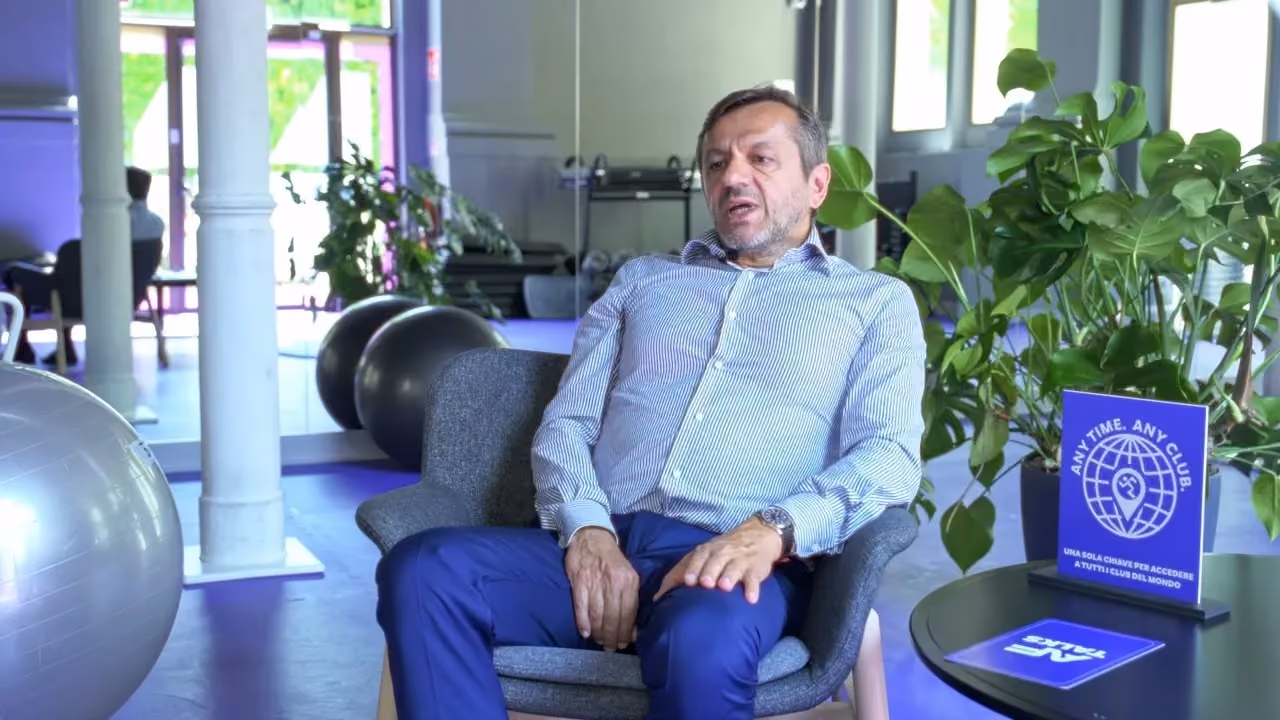 Un uomo in camicia a righe è seduto su una poltrona grigia durante un'intervista in uno studio Anytime Fitness. Sul fondo si vedono palle da ginnastica, attrezzature per l'allenamento e piante da interno.