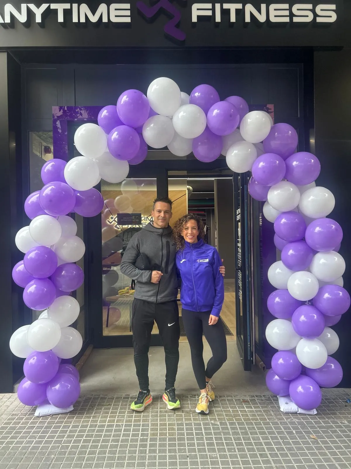 Hombre y mujer de pie bajo un arco de globos morados y blancos frente a la entrada de un gimnasio Anytime Fitness, sonriendo y vestidos con ropa deportiva.