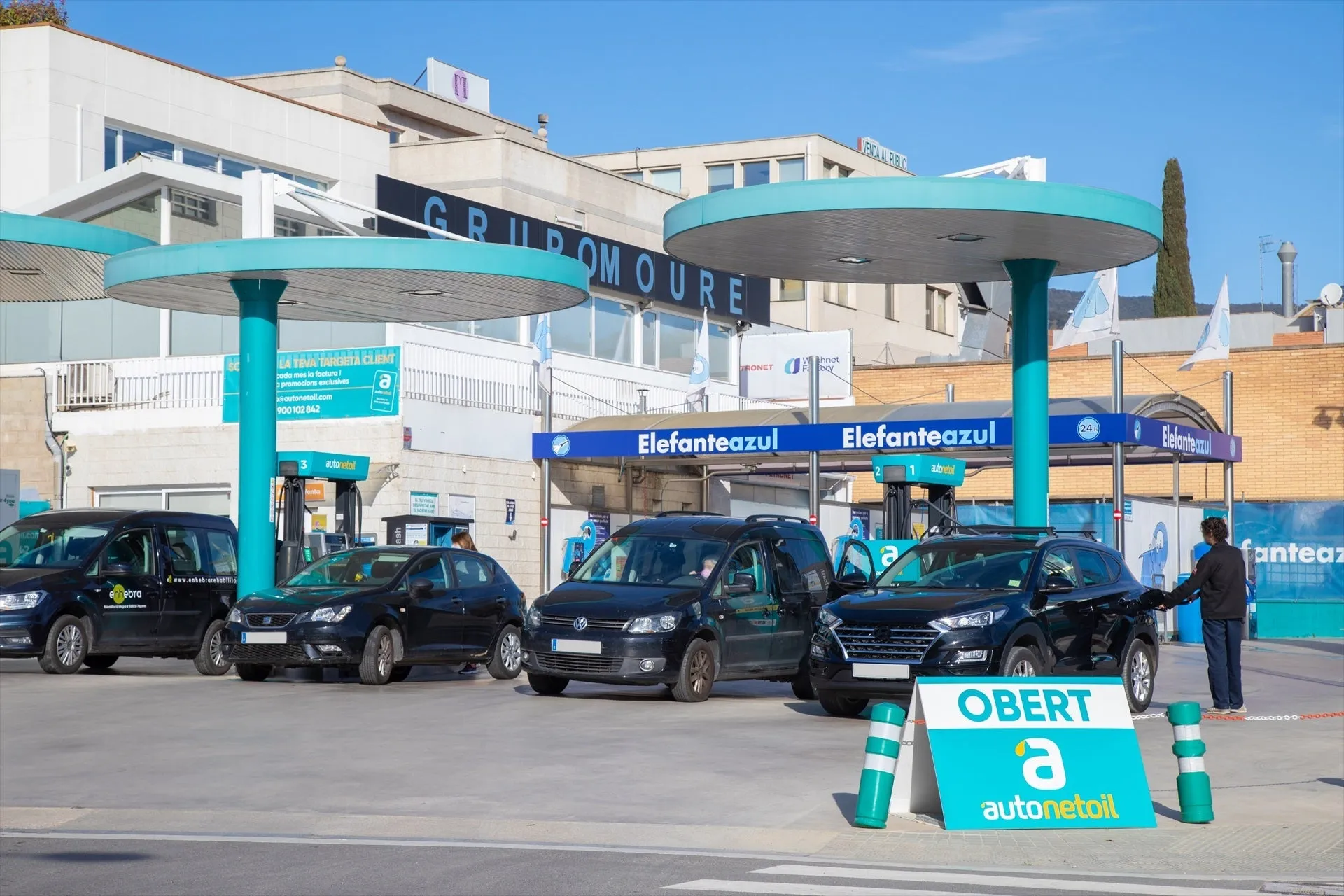 Gasolinera llamada Elefanteazul con tres coches negros repostando bajo toldos circulares de color verde azulado en un día soleado.
