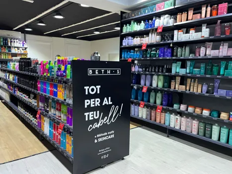 Pasillo de la tienda con estantes repletos de coloridos productos para el cuidado del cabello y la piel y un letrero negro en el centro que dice «Beth's Tot per al teu cabell! Método curly & skincare.