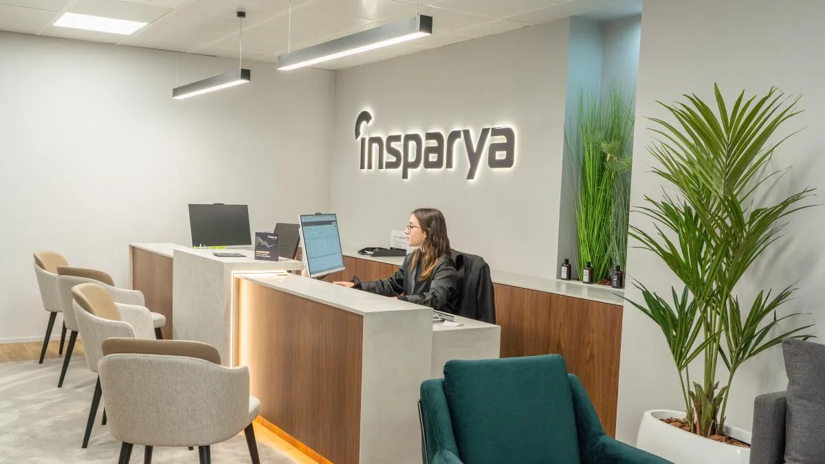 Moderna recepción de oficina con una mujer trabajando en una computadora detrás de un escritorio blanco y de madera, sillas de color beige para los visitantes, plantas verdes y un letrero en la pared que dice «insparya».